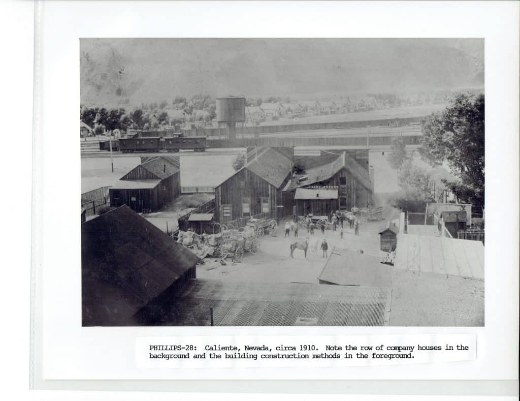 A black and white photo shows a dusty town with wooden buildings, people, horses, and a train in the distance.