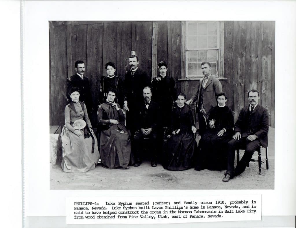 A black and white photo shows a family of ten, men in suits and women in long dresses, posing in front of a wooden building.