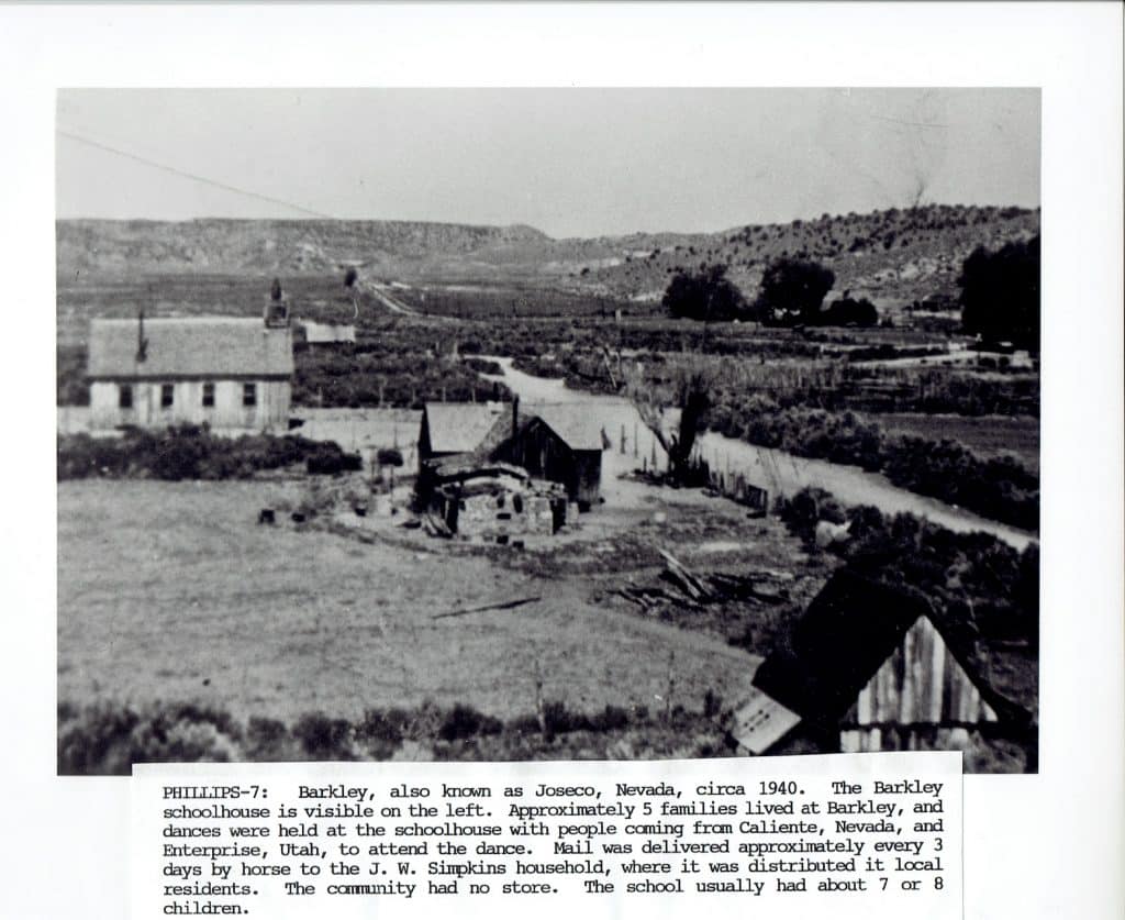 A black and white photo shows a small schoolhouse, other buildings, and a road in a rural valley.