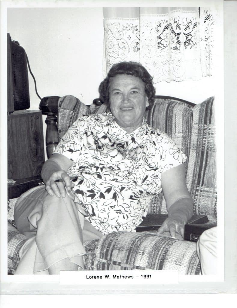 A smiling woman with dark curly hair sits in a patterned armchair, wearing a floral shirt and light pants.
