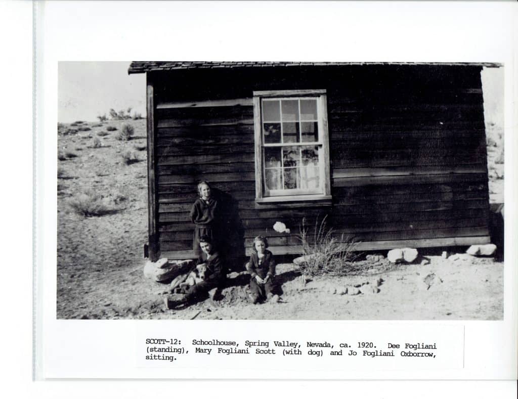 A black and white photo shows a woman standing and two children sitting outside a wooden schoolhouse.