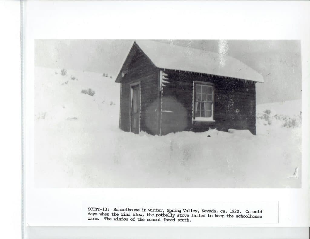 A black and white photo shows a small, snow-covered wooden schoolhouse with a single window and door.