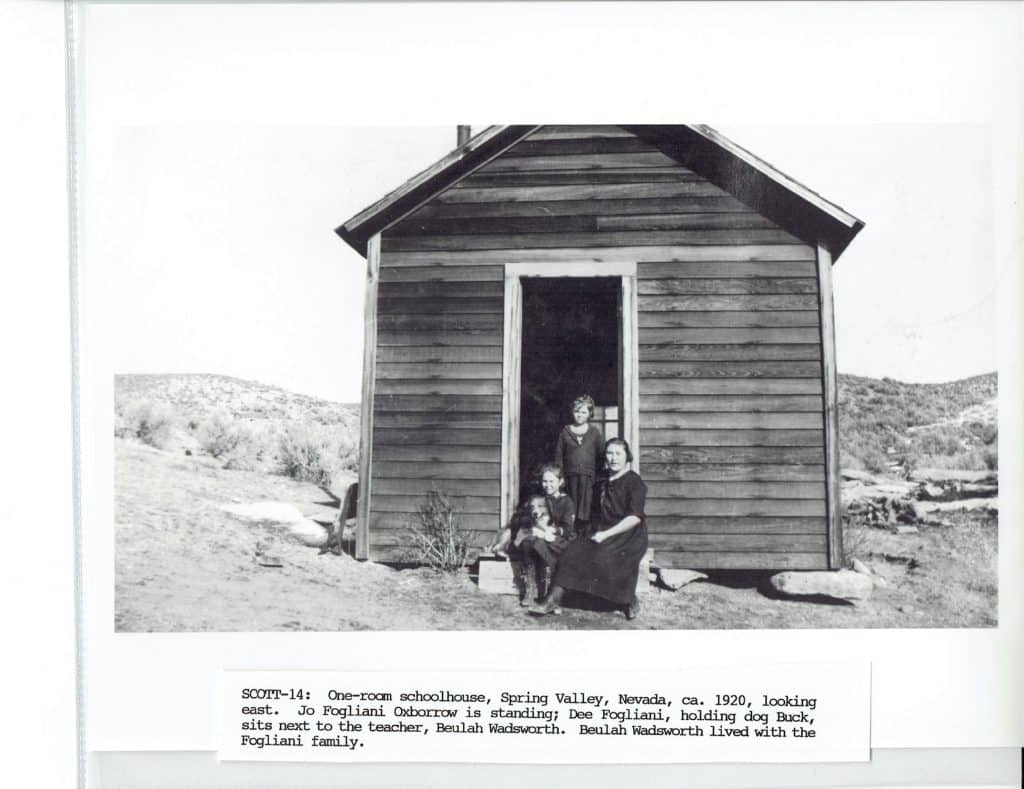 A black and white photo of three people and a dog in front of a wooden schoolhouse.