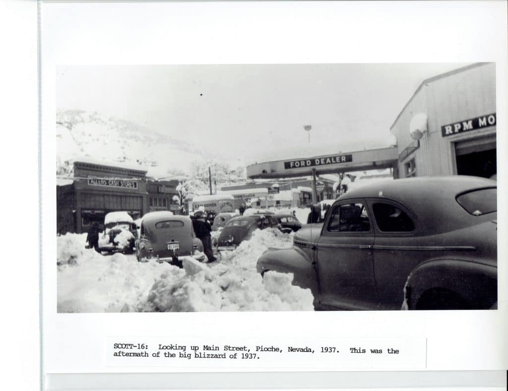 Black and white photo of a snow-covered street with cars and buildings, including a "FORD DEALER" sign.