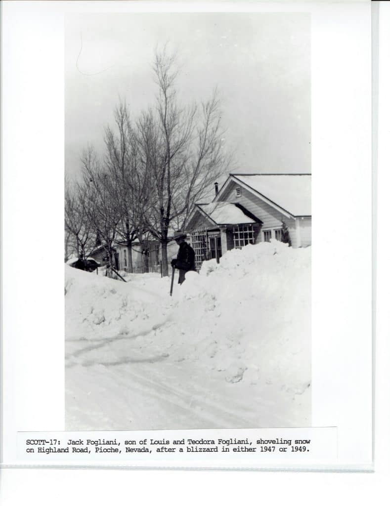 A man shovels snow from a street with towering snowdrifts, bare trees, and houses in the background.