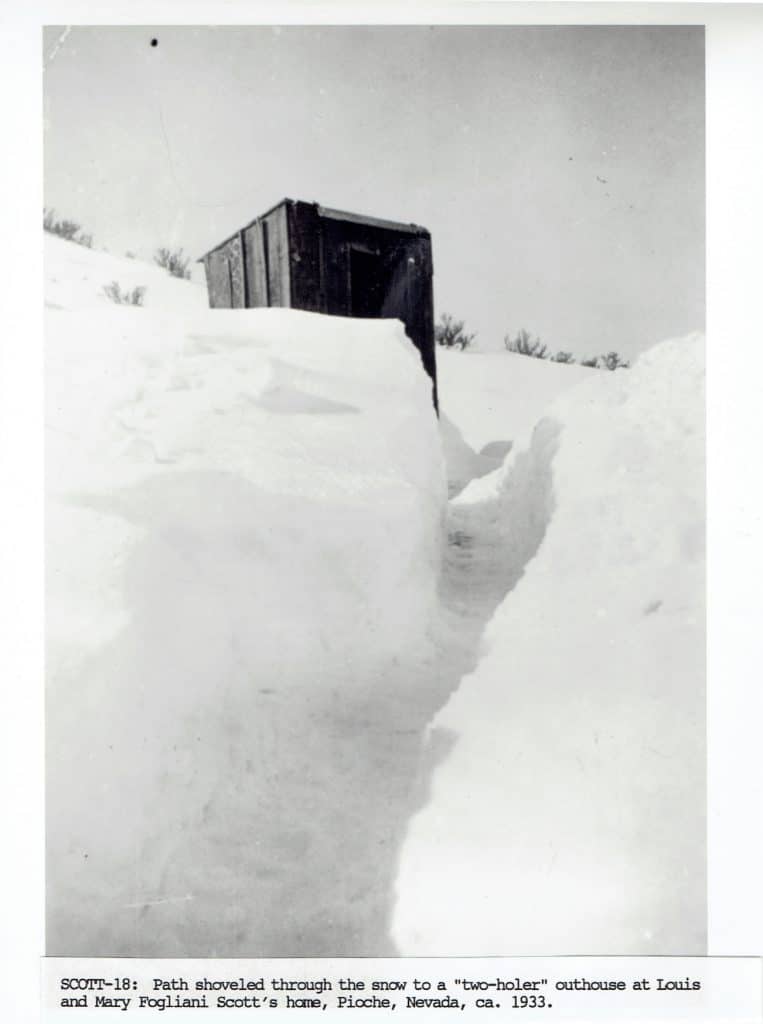 A narrow path shoveled through deep snow leads to a small, dark wooden outhouse under a bright sky.
