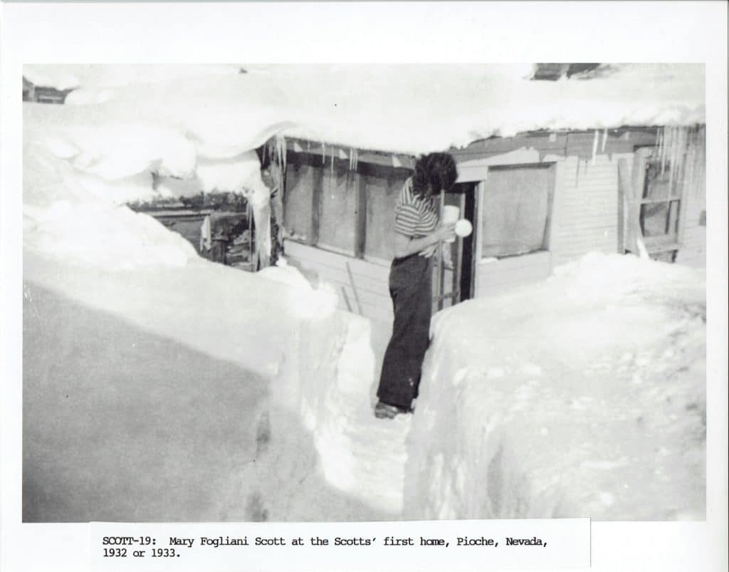 A person stands in a deep, snow-filled trench, holding a snowball near a house with icicles.