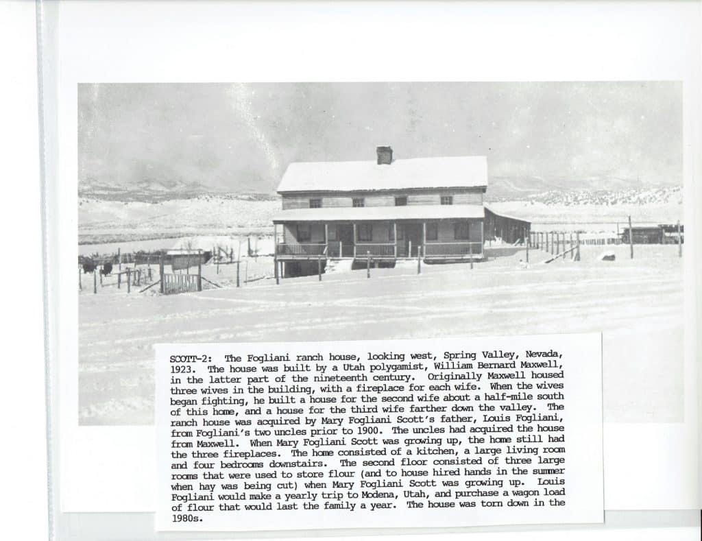 A black and white photo of a two-story house with a porch, surrounded by snow and mountains.