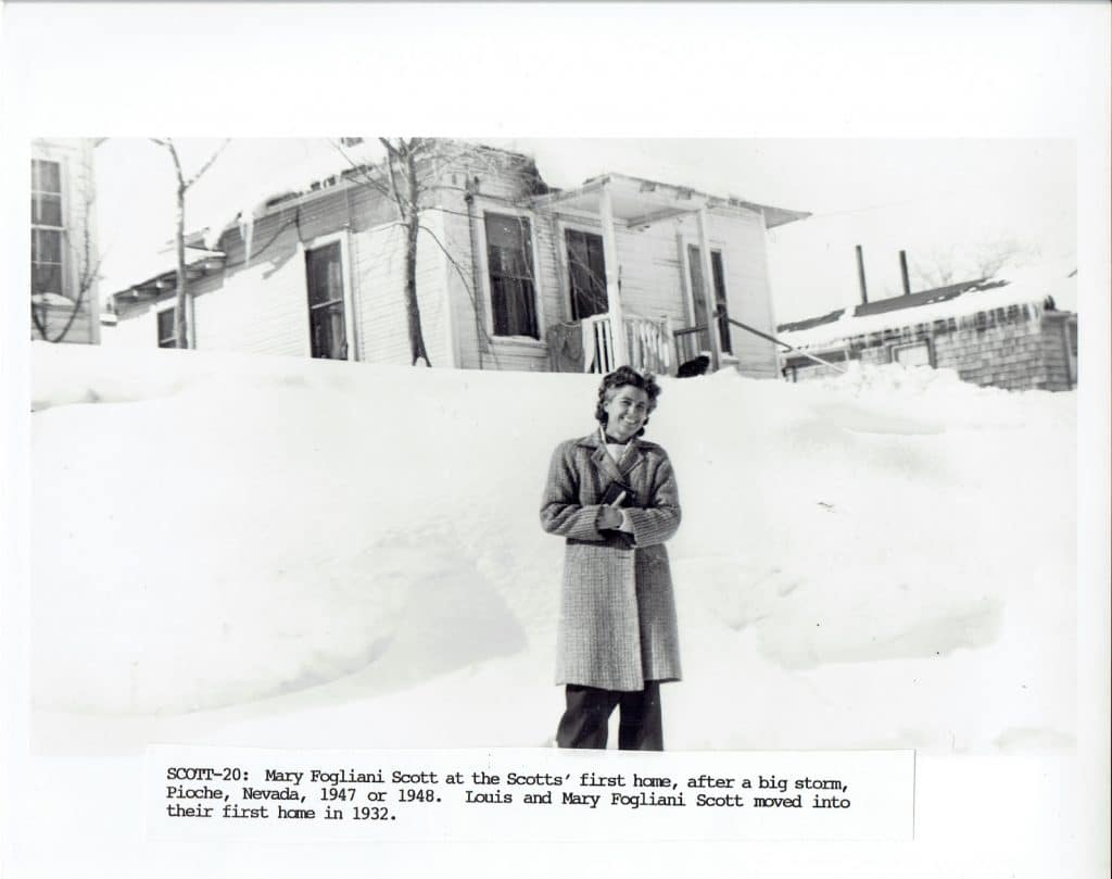 A smiling woman in a coat stands in deep snow before a house, with text "SCOTT-20" below.
