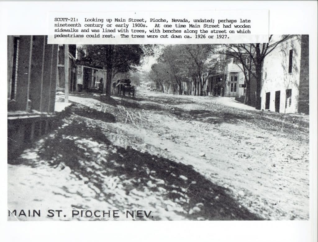 A black and white photo of a dirt road with buildings and trees on either side, labeled "MAIN ST. PIOCHE NEV."
