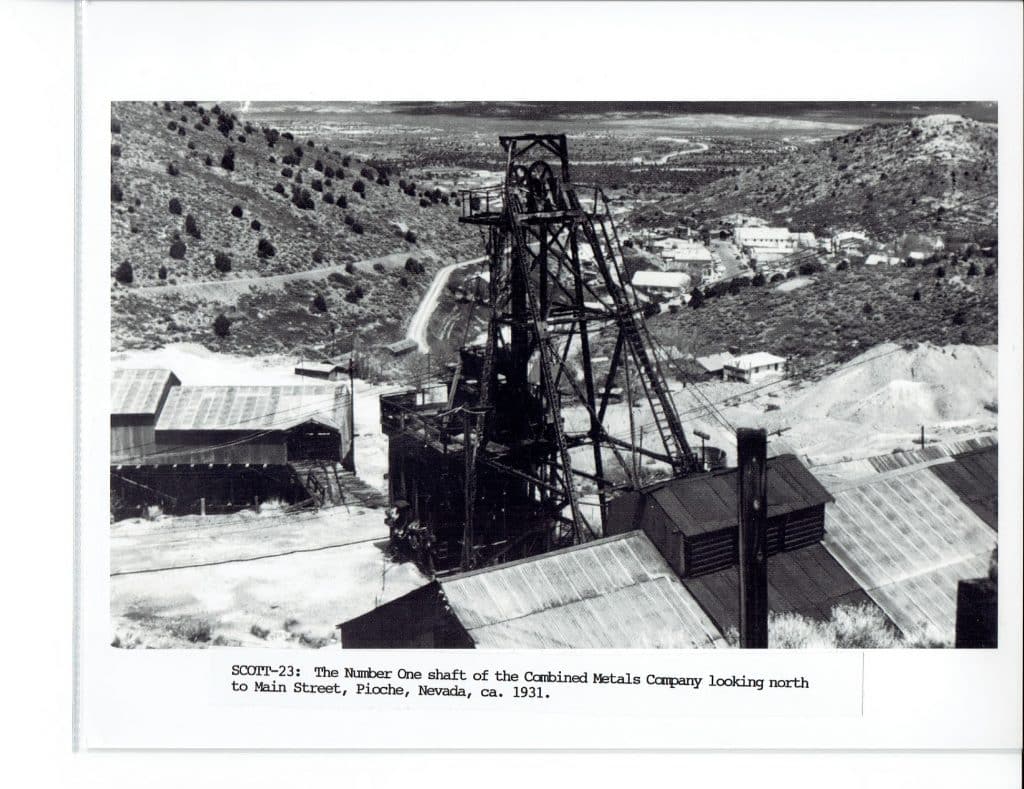 A black and white photo shows a large mining headframe and buildings on a hillside overlooking a town.