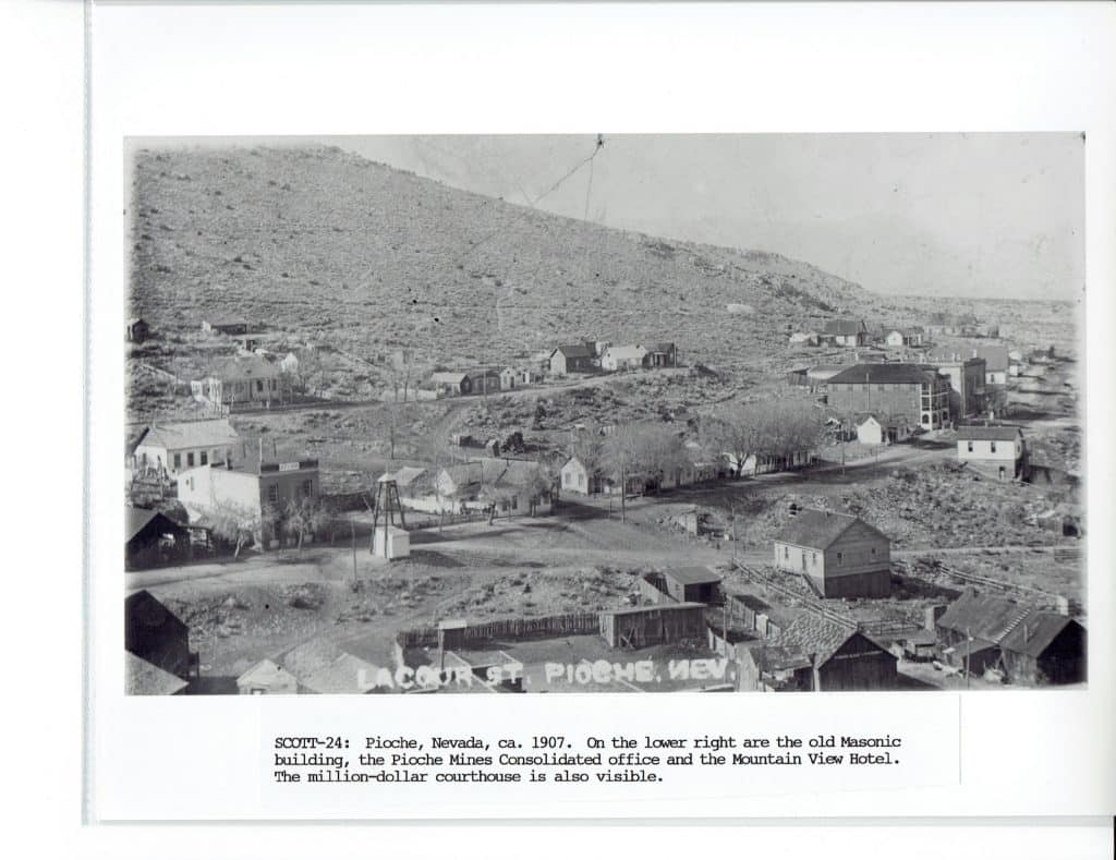 A black and white photo shows a small town nestled in a valley with a large, barren hill behind it.
