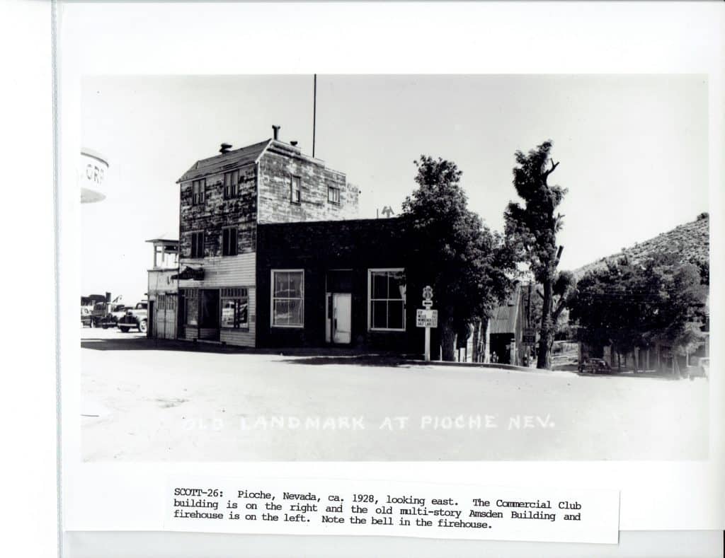 A black and white photo shows two old buildings, trees, and a distant hill under a bright sky.