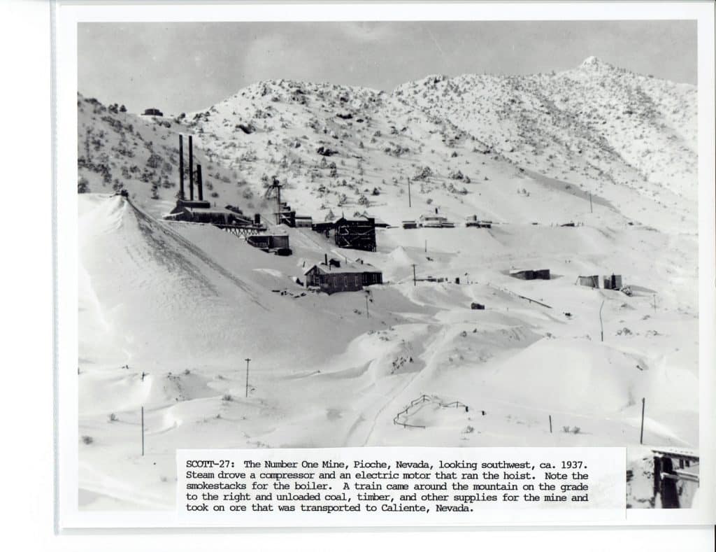 A black and white photo shows a snowy mountain with several buildings, including three tall smokestacks.