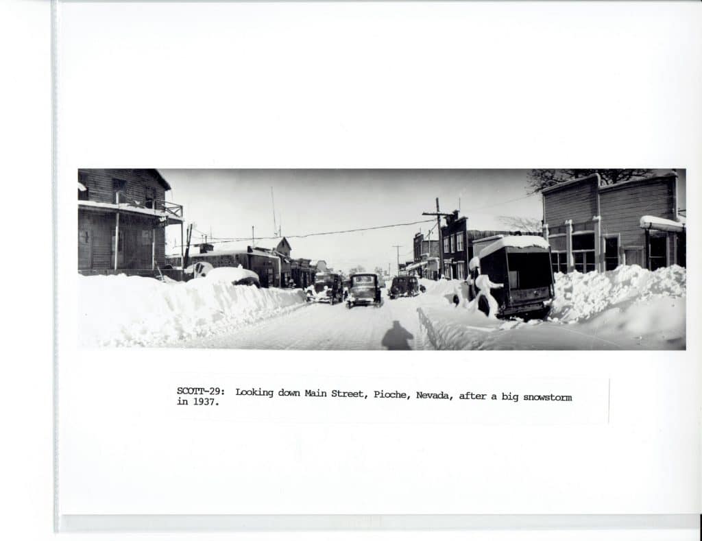 Black and white panoramic photo of a snow-covered street with buildings and cars, "SCOTT-29: Looking down Main Street, Pioche, Nevada, after a big snowstorm in 1937."