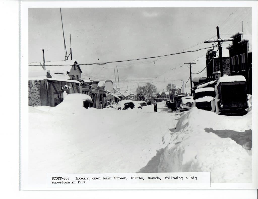 A black and white photo of a snow-covered street with buildings and cars buried in deep snow.