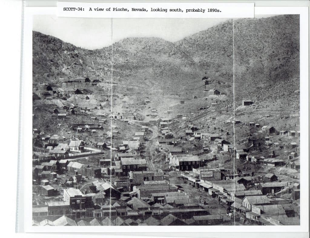 A black and white photo shows a mining town with many buildings nestled in a valley between hills.