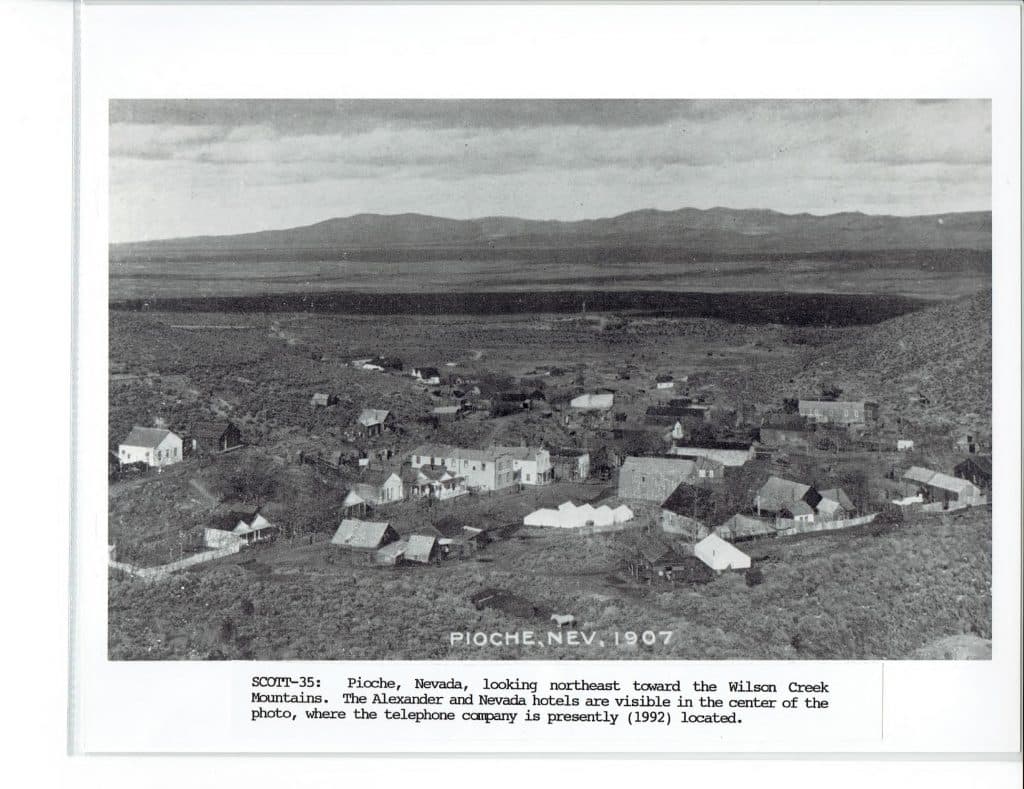A black and white aerial photo shows a small town with many buildings nestled in a vast, arid landscape, with "PIOCHE, NEV. 1907" at the bottom.
