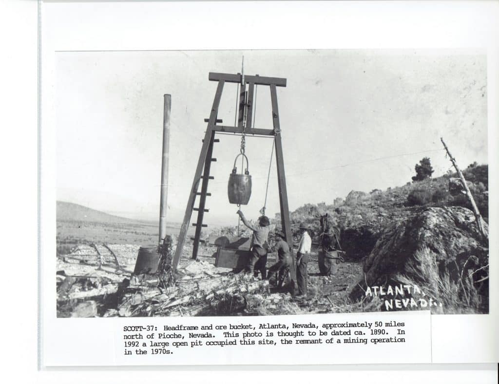 Three men operate a headframe and ore bucket in a rocky, barren landscape with "ATLANTA NEVADA" on a rock.