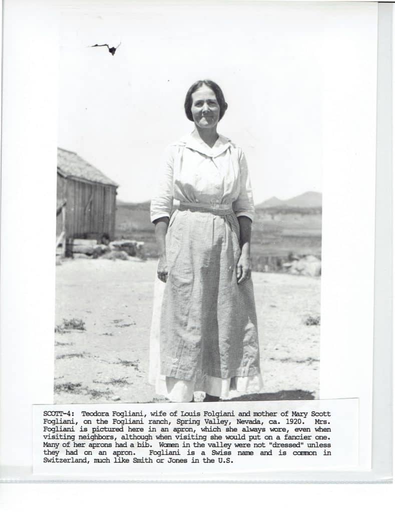 A smiling woman in a long apron and dress stands outdoors with a wooden shack behind her.