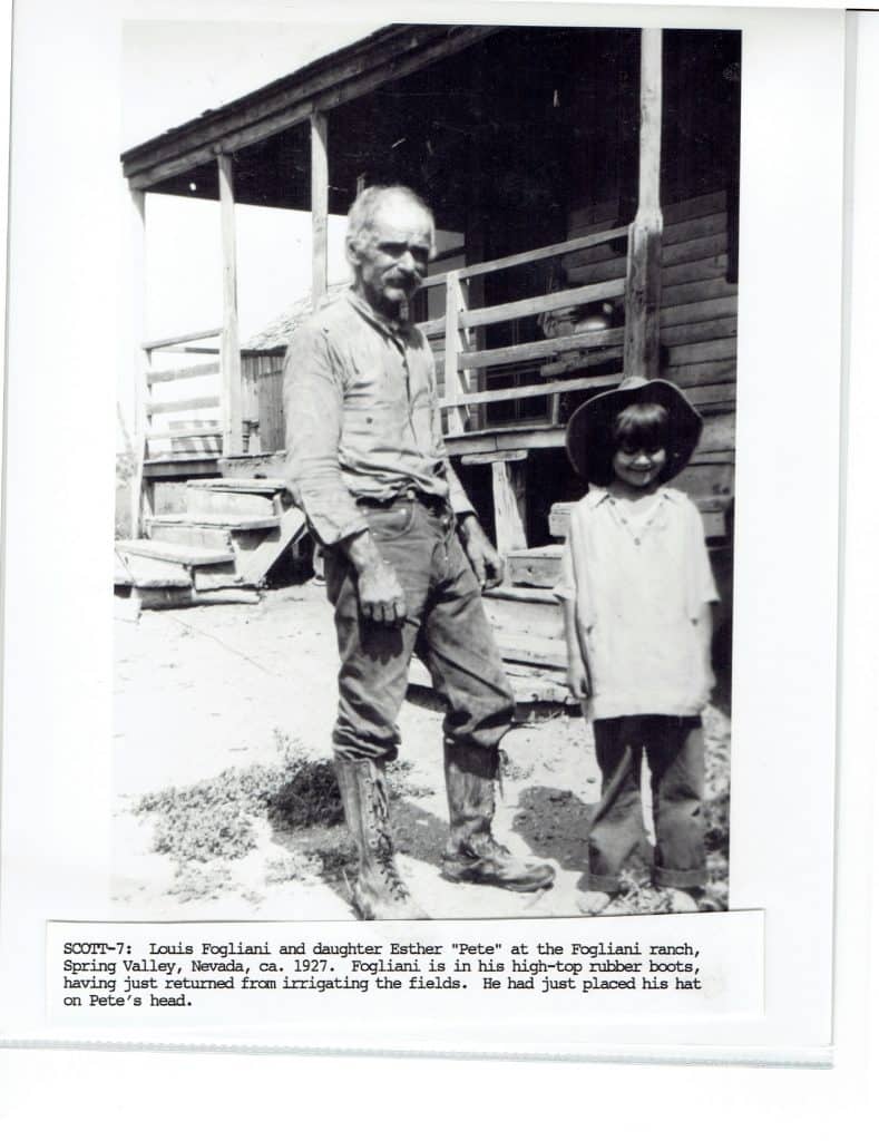 A man in work boots and a child in a hat stand outside a wooden building with a porch.