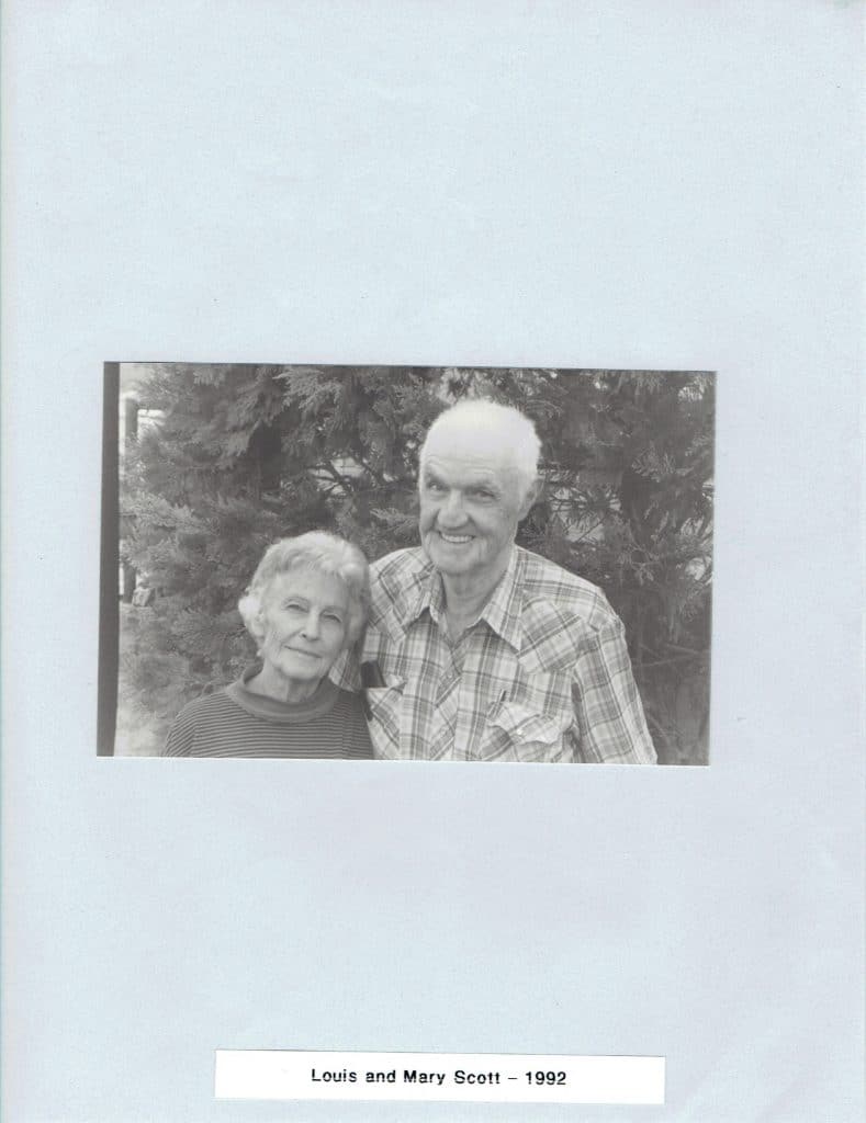 A smiling elderly couple, a woman in a striped shirt and a man in a plaid shirt, stand before trees. "Louis and Mary Scott - 1992" is below.