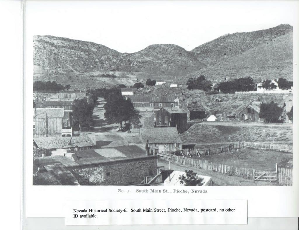 Black and white photo of a town with scattered buildings and trees, nestled in a valley between large hills, with text "No. 5. South Main St., Pioche, Nevada".