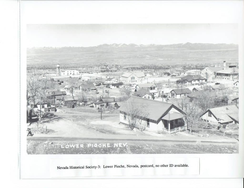 Black and white photo of a town with many houses, a water tower, and mountains in the background.