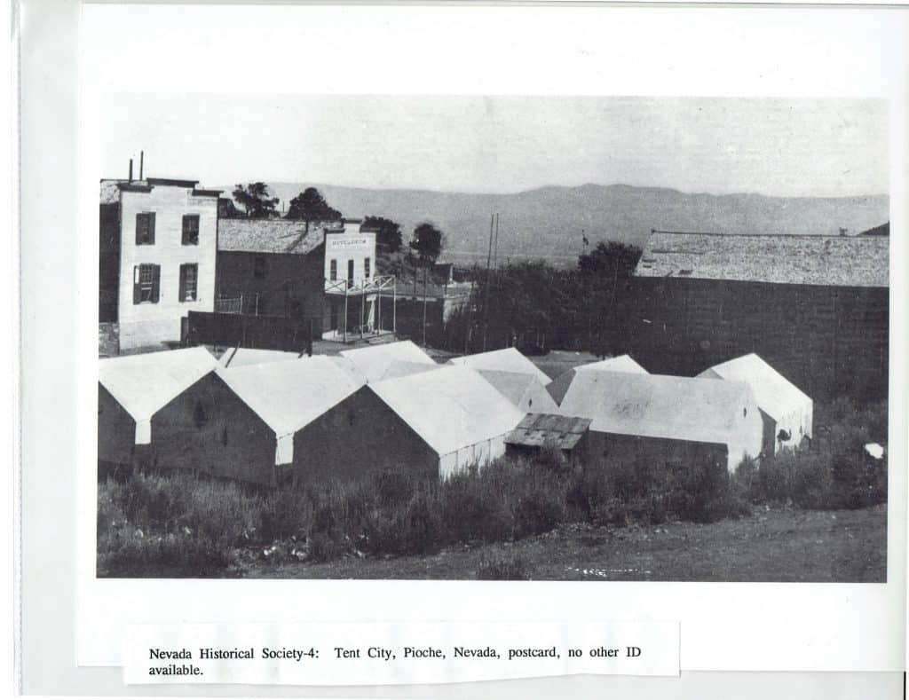 Black and white photo of a tent city with several canvas tents in the foreground and buildings behind them.