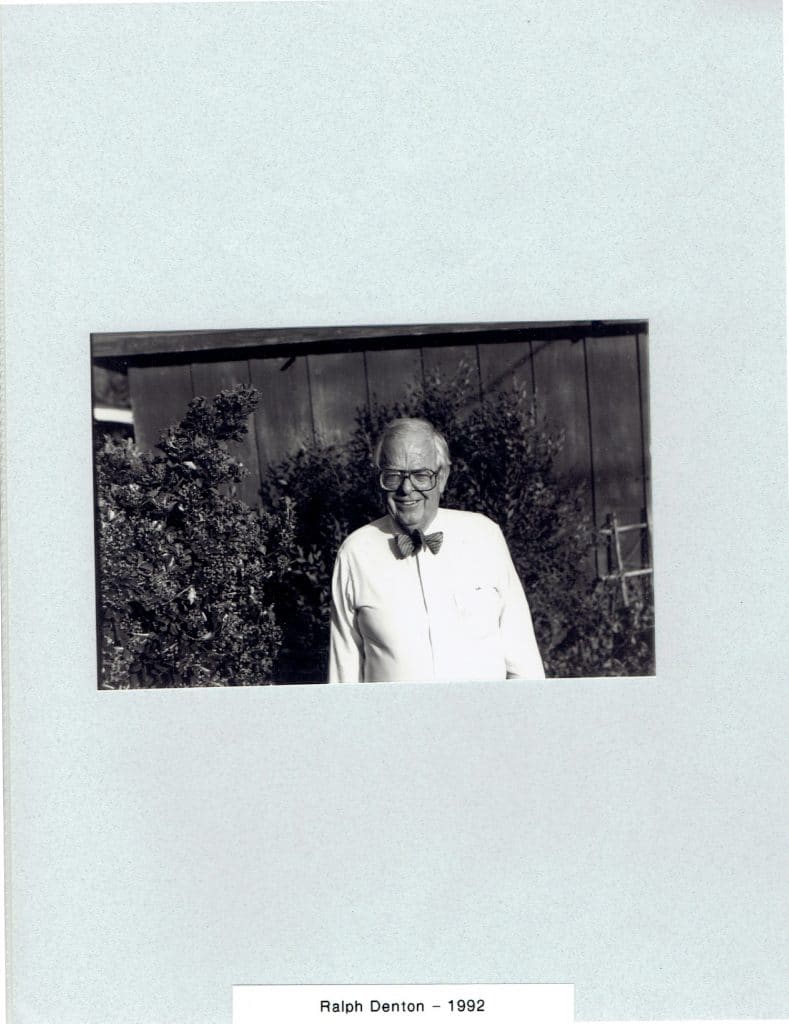 A smiling older man with glasses and a bow tie stands in front of bushes and a building. "Ralph Denton - 1992"