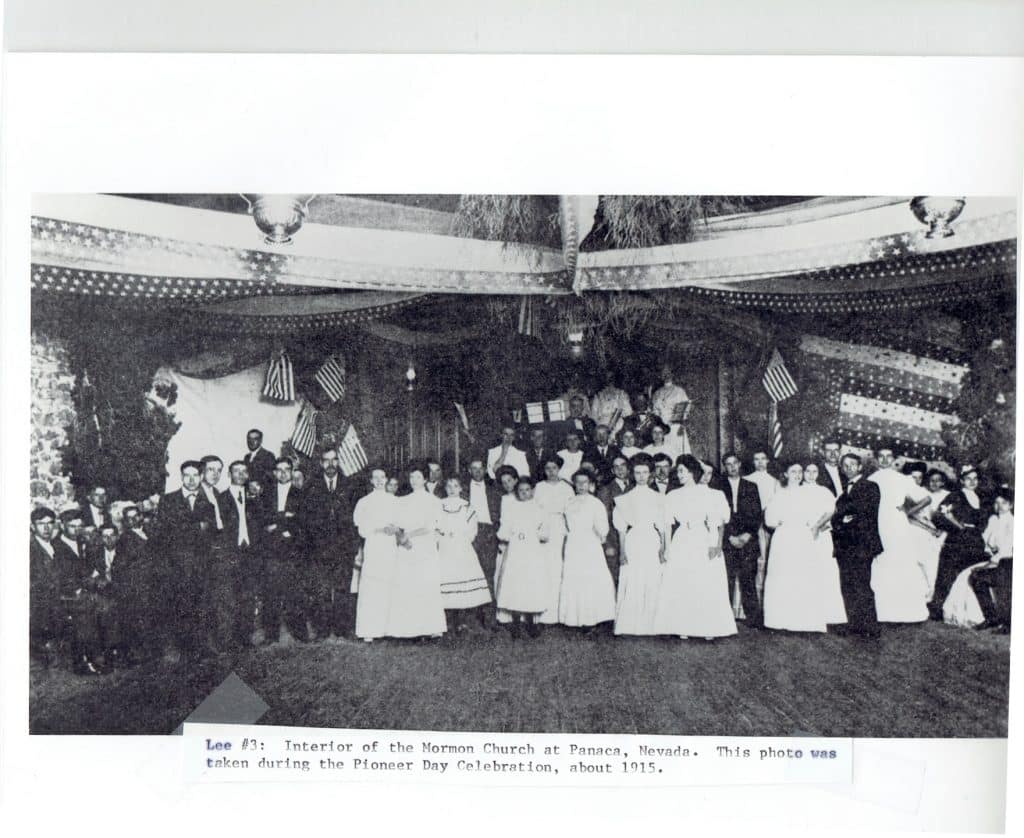 A large group of people, men in suits and women in white dresses, stand in a decorated hall.