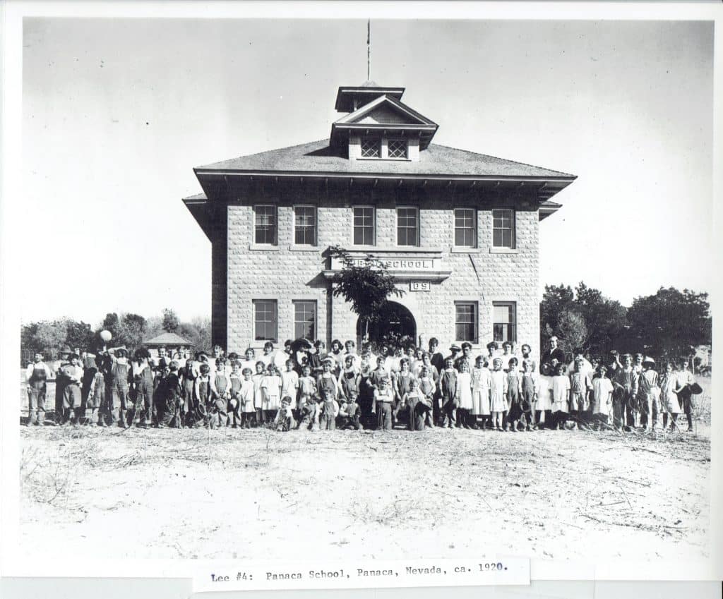 A large group of children and adults stand in front of a stone building with "PUBLIC SCHOOL" above the entrance.