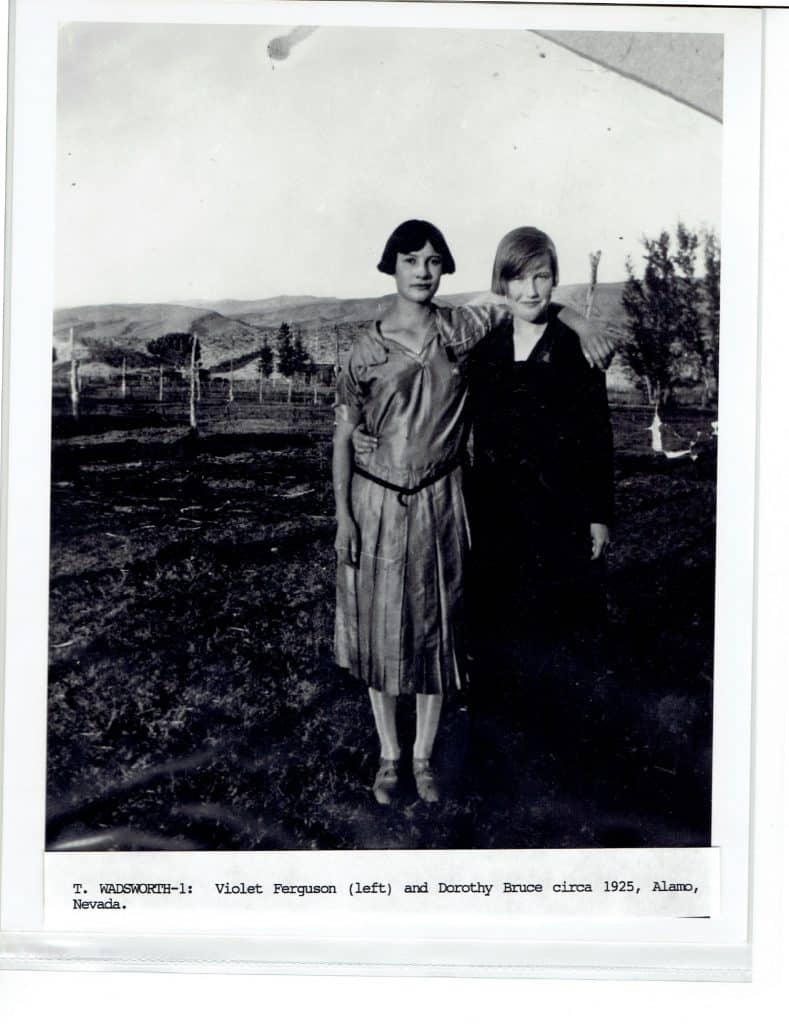 Two young women, one in a light dress and one in a dark dress, stand with arms around each other outdoors.