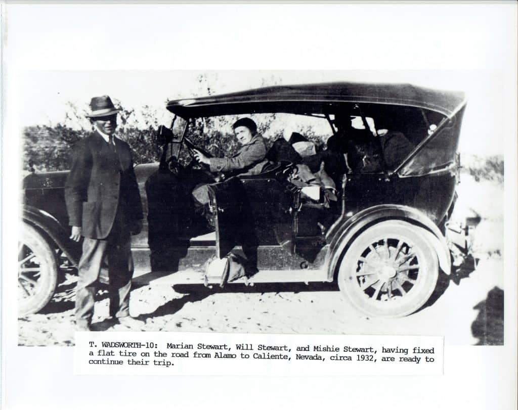 A man in a suit stands beside a vintage car with a woman sitting in the driver's seat, "T. WADSWORTH-10".