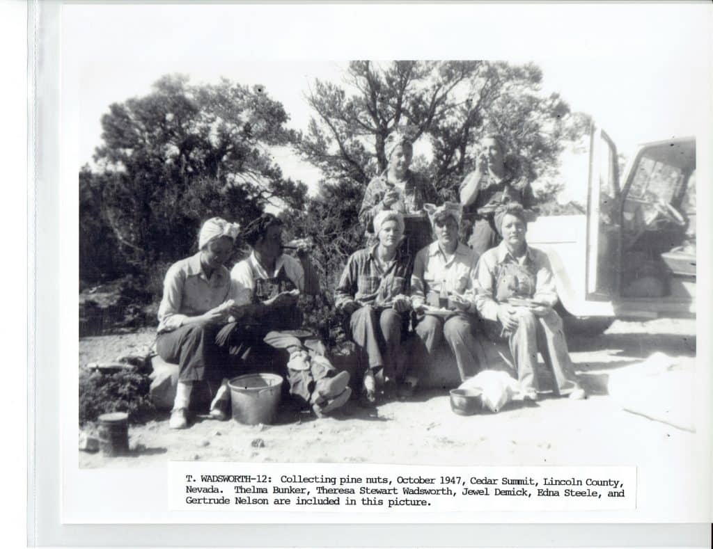 Seven women in head coverings and work clothes sit outdoors with buckets and a truck.