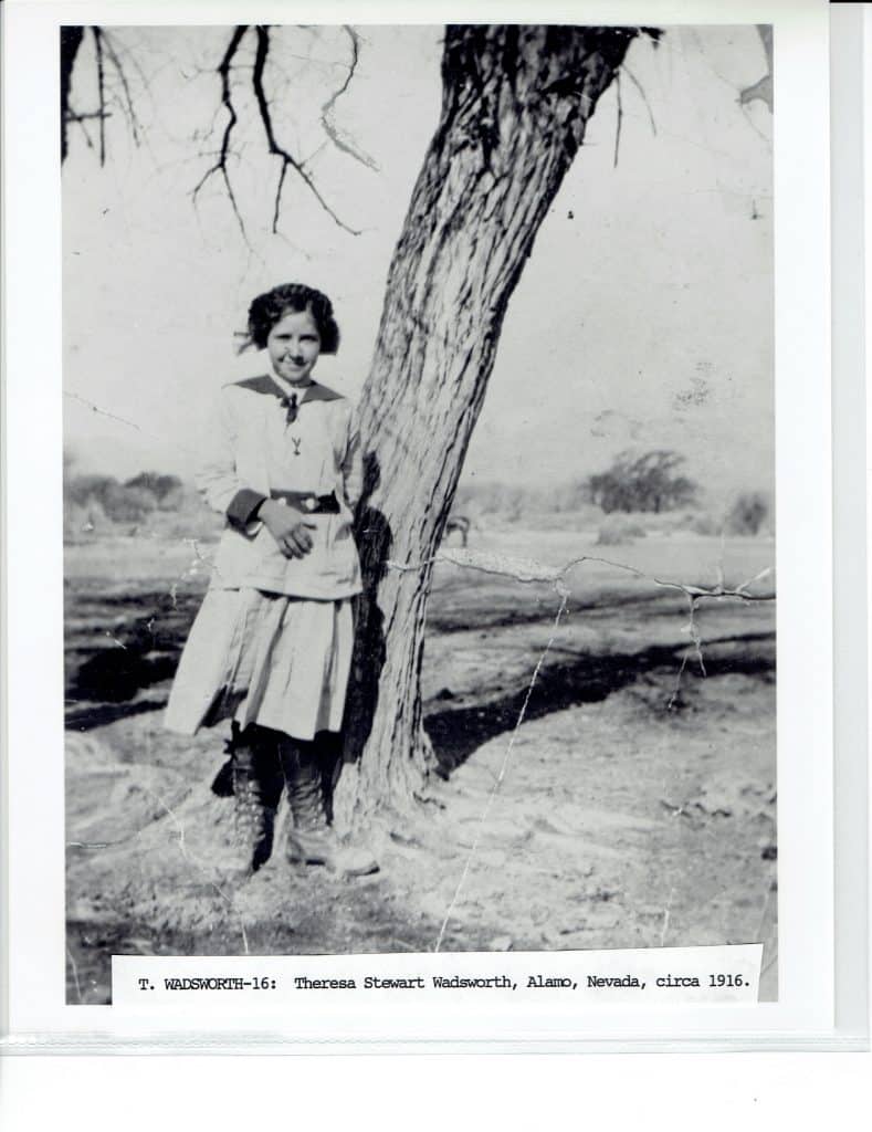 A young woman in a light skirt and jacket stands smiling next to a tree in a desert landscape.