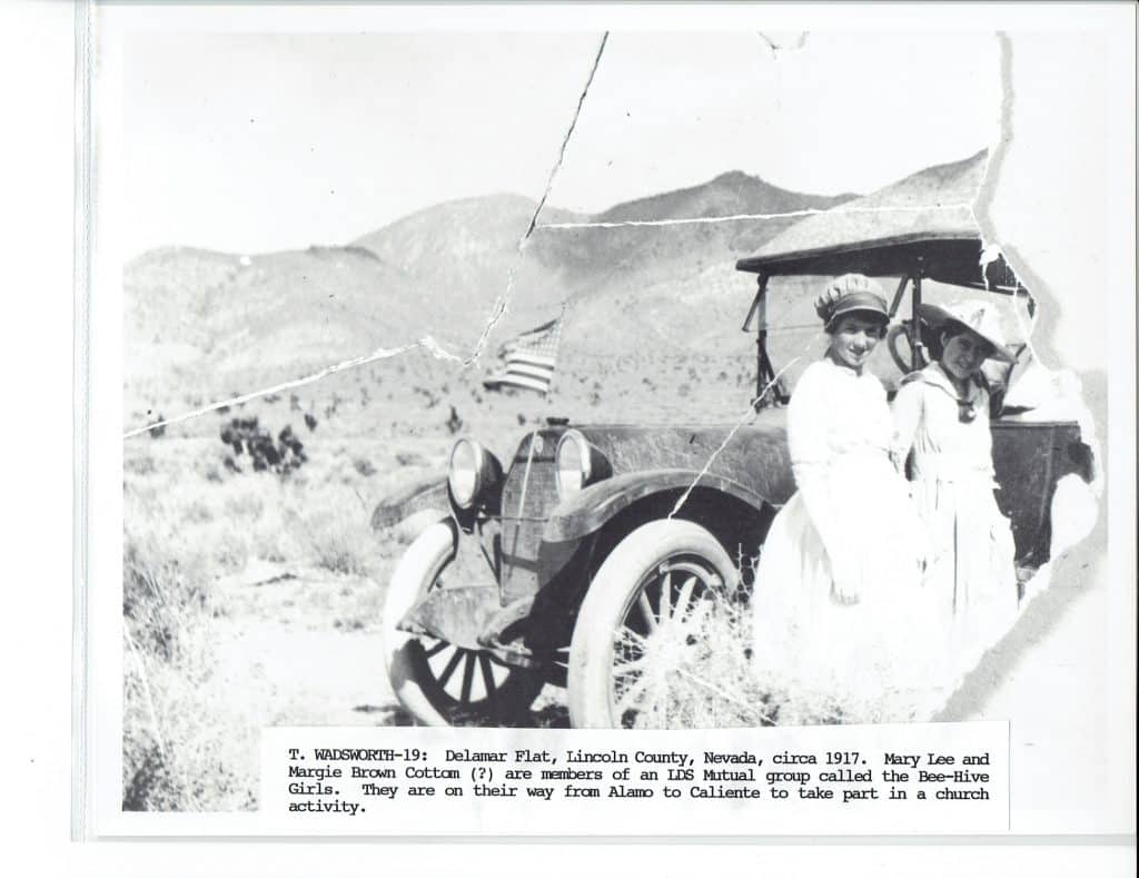 Two young women in hats and long dresses stand beside an old car with an American flag, in a desert landscape.