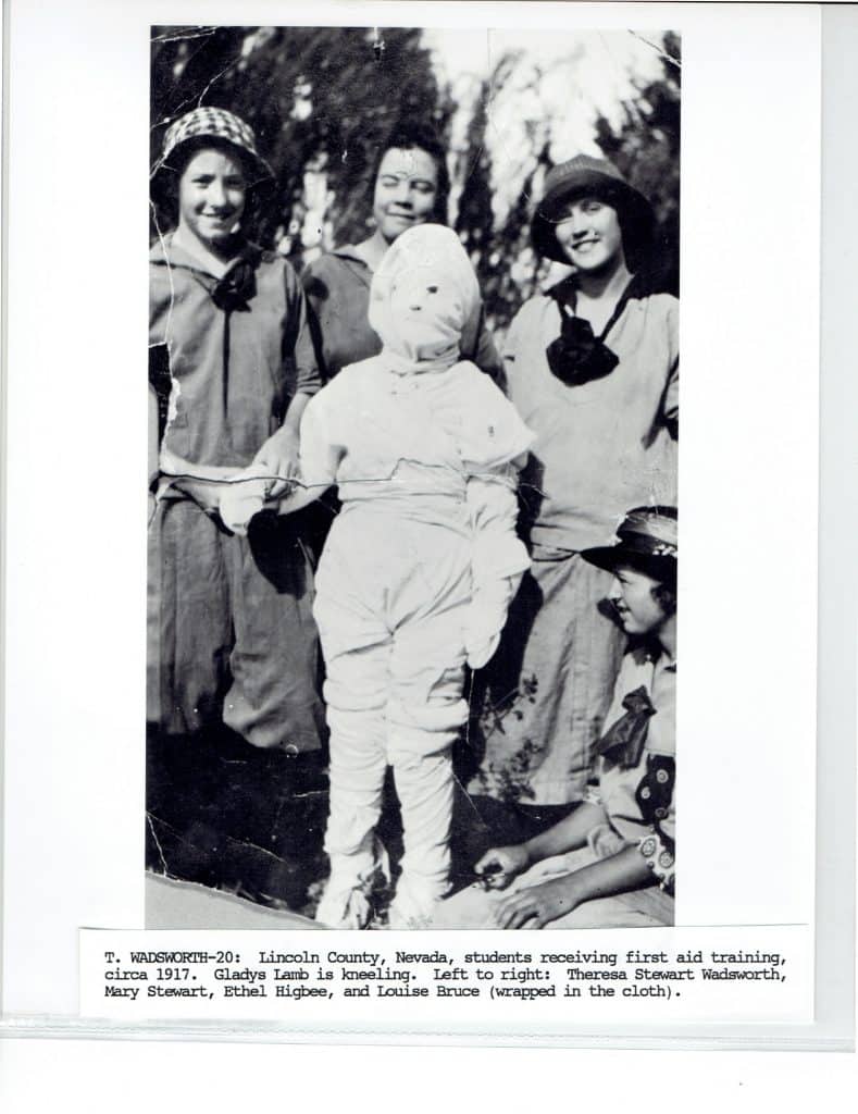 Five smiling students in hats and dresses, one wrapped in cloth, pose outdoors for first aid training.