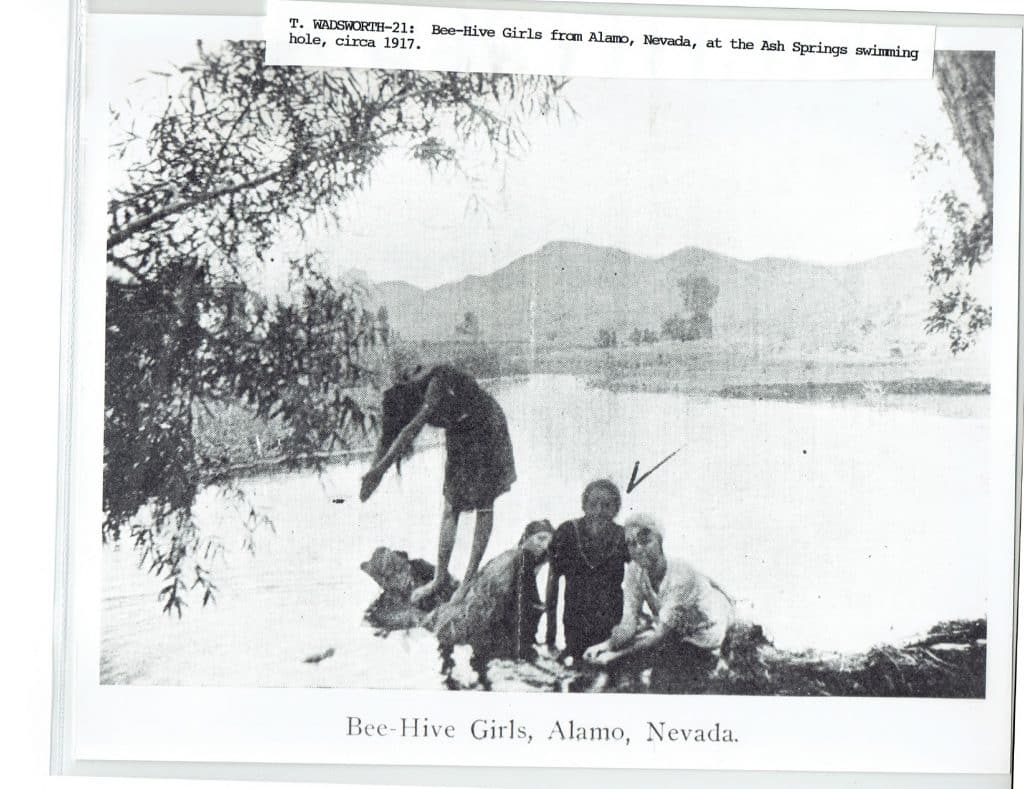 Four girls in dark clothing are by a swimming hole with mountains in the background, "Bee-Hive Girls, Alamo, Nevada."