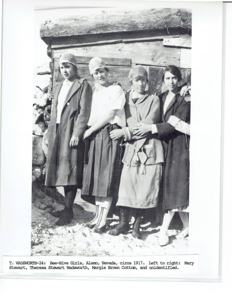 Four young women in long coats and bonnets stand in front of a wooden structure.