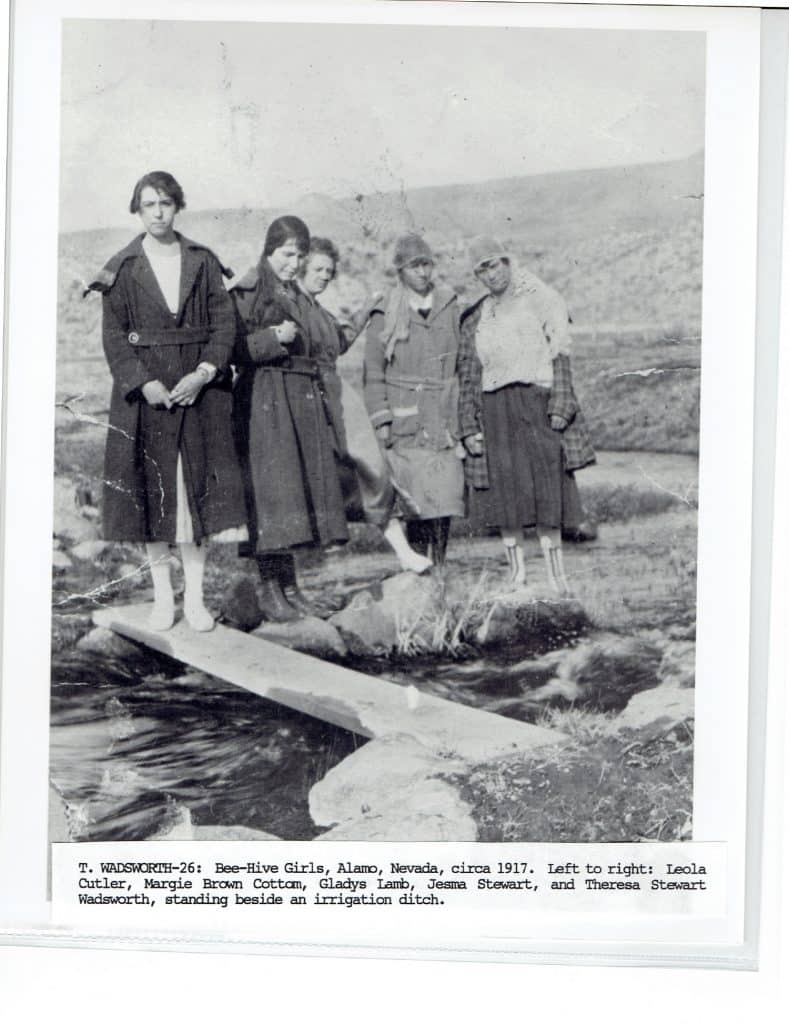 Five young women in coats and long skirts stand by an irrigation ditch in a desert landscape.