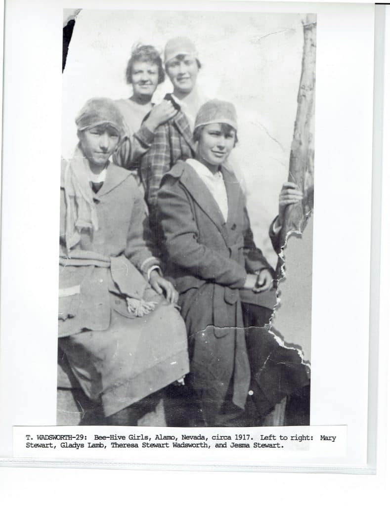 Four young women in hats and coats, two standing and two seated, pose outdoors in a faded black and white photo.