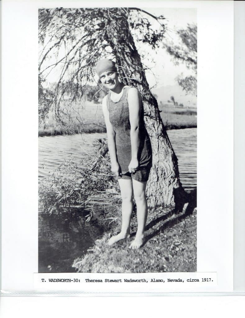 A smiling woman in a dark swimsuit and cap stands barefoot by a tree and water.