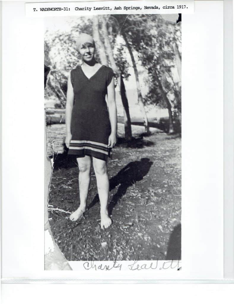 A smiling woman in a dark swimsuit and cap stands barefoot on grass, with trees in the background.