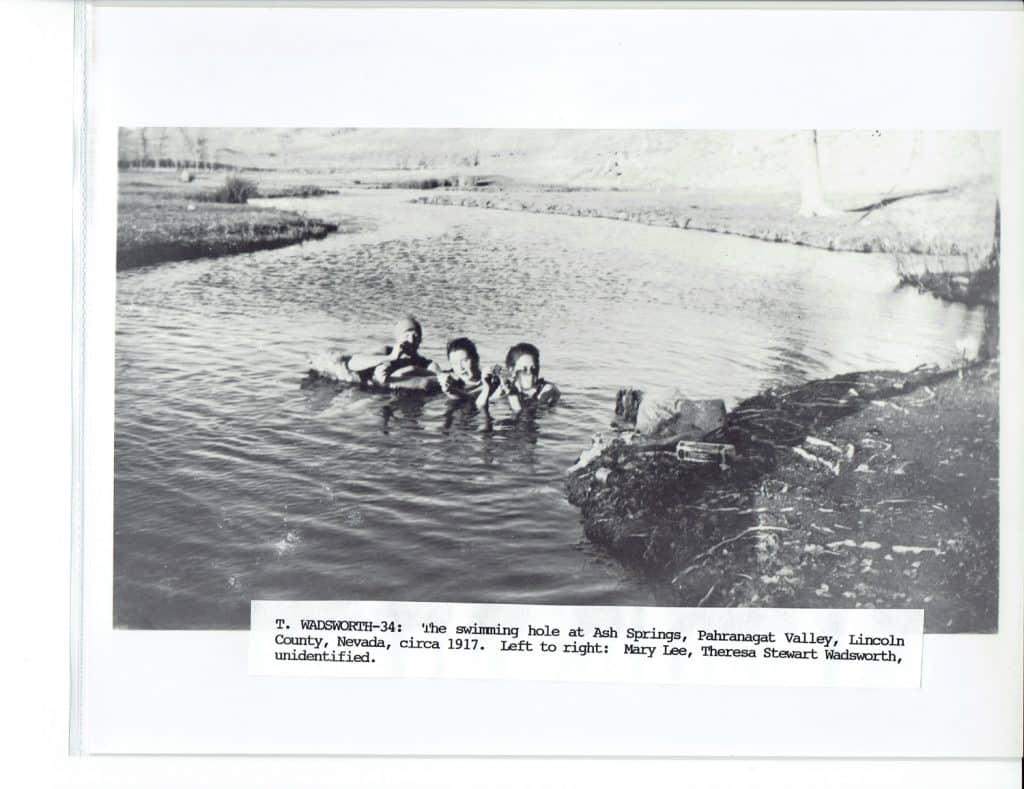 Three people swim in a river with grassy banks, with text: "T. WADSWORTH-34: the swimming hole at Ash Springs, Pahranagat Valley, Lincoln County, Nevada, circa 1917. Left to right: Mary Lee, Theresa Stewart Wadsworth, unidentified."