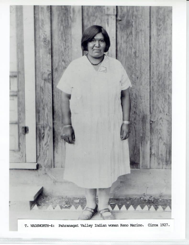 A black and white photo of a woman with short dark hair, wearing a light dress, standing against a wooden wall.