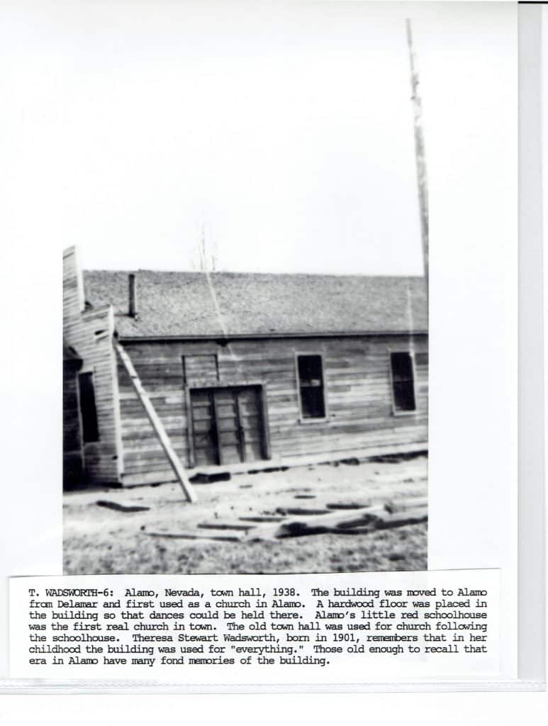A black and white photo shows a weathered wooden building with a slanted roof and two windows.