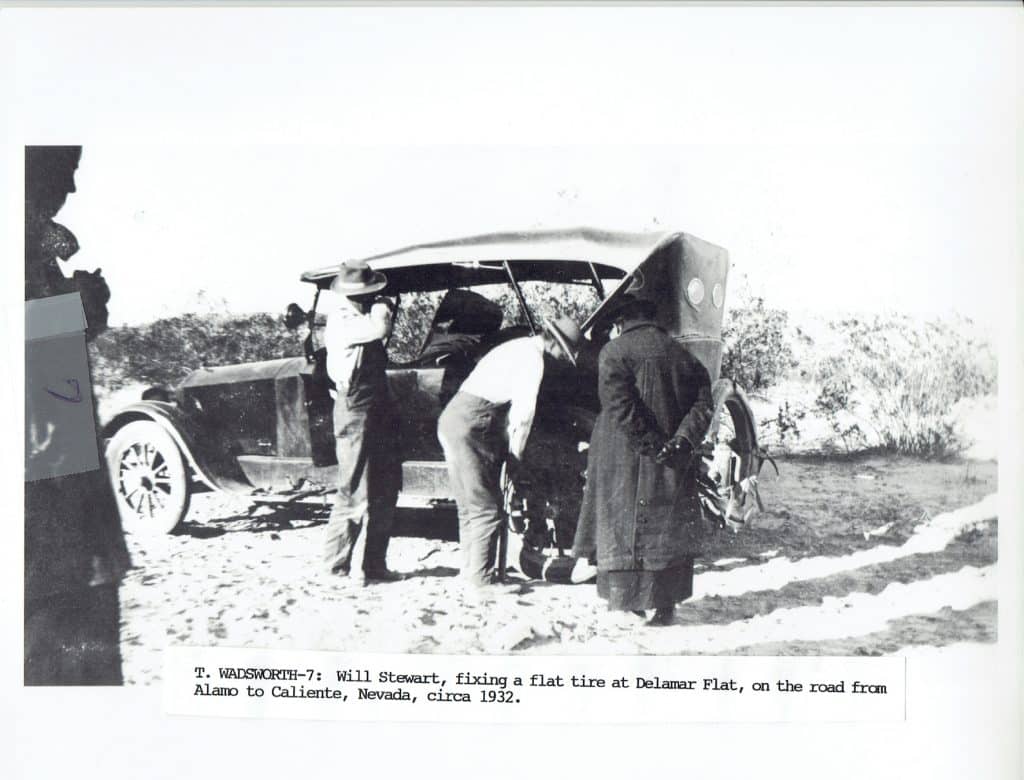 Three people are gathered around a vintage car with its hood open on a dirt road.