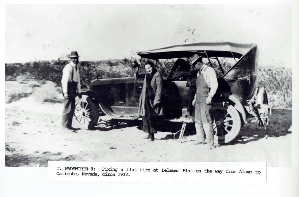 Three people stand around an old car with an open trunk and a flat tire in a desert landscape.