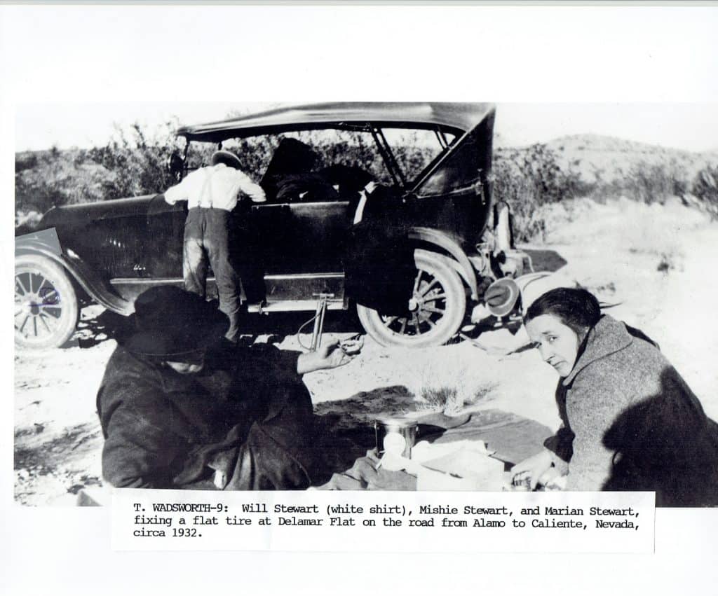 A man in a white shirt fixes a flat tire on a vintage car while two women sit on the ground.